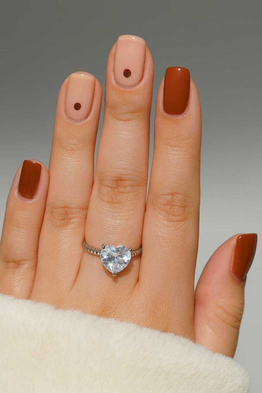 A hand with a heart-shaped diamond ring wears a manicure with brown and nude nails. The nude nails have a small brown dot near the cuticle, and the person is wearing a white fuzzy sleeve.