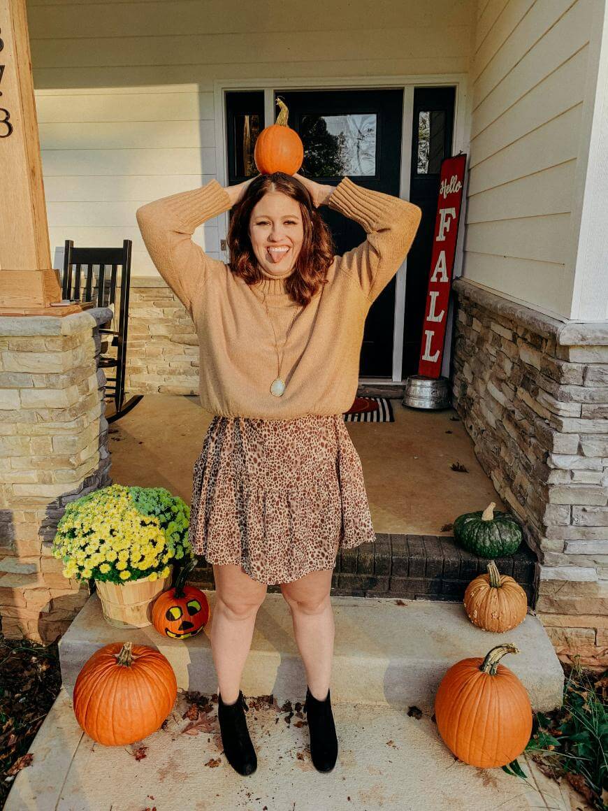 A woman stands on a porch decorated with pumpkins and fall flowers, playfully holding a pumpkin on her head and sticking out her tongue. She wears a tan sweater, patterned skirt, and black boots. A “Hello Fall” sign is by the door.