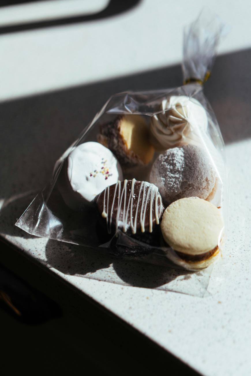 A clear plastic bag filled with assorted cookies and pastries, including macarons and chocolate-dipped treats, sits on a sunlit countertop.
