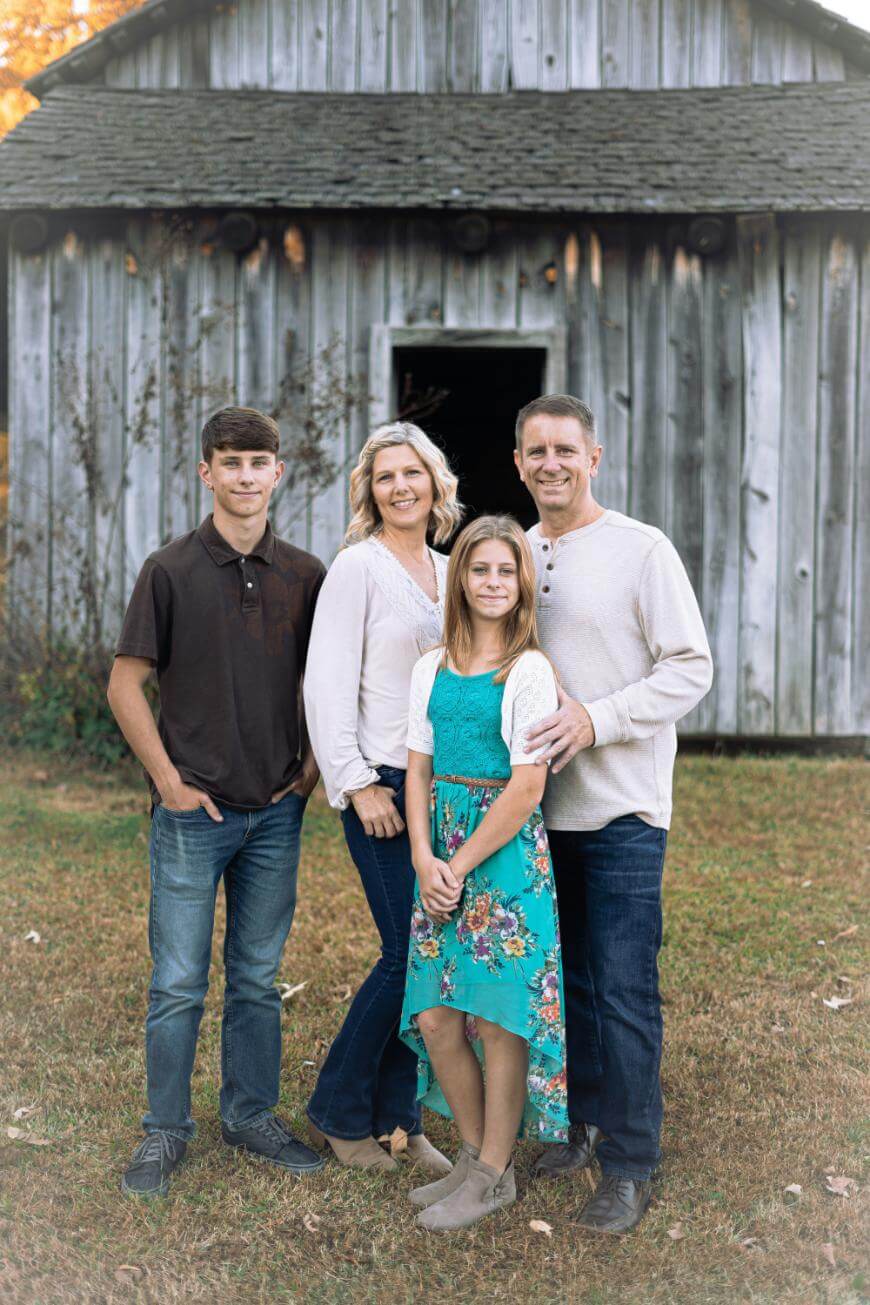 A family of four poses in front of a rustic wooden barn. Two adults stand behind a teenage boy and girl. They all smile, dressed in casual fall clothing, standing on grass with the weathered barn as the backdrop.