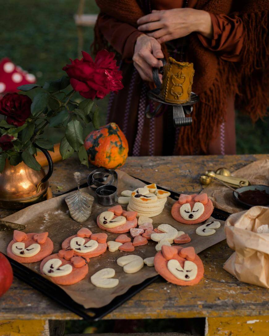 A person prepares fox-shaped cookies on a rustic table decorated with roses, a gourd, and baking tools. Some cookies are on a tray, while others are being decorated. Warm colors and cozy atmosphere suggest autumn or a festive occasion.