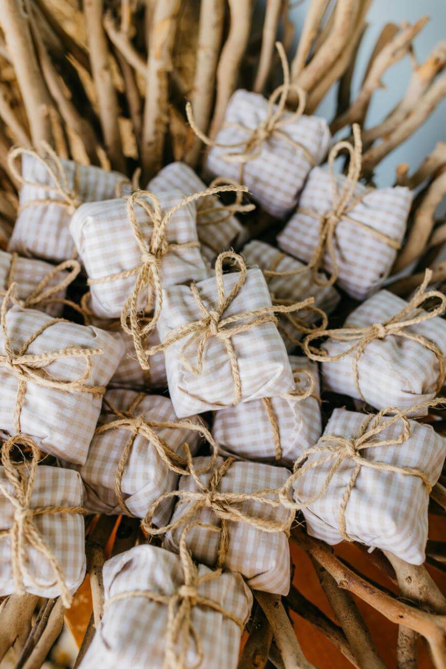 Small fabric-wrapped gifts tied with twine are stacked together on a pile of light-colored wooden branches, creating a rustic and charming display.
