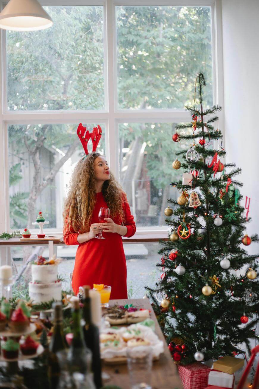A woman in a red dress and reindeer antlers stands by a decorated Christmas tree, holding a glass of wine. A table with festive food and drinks is in the foreground, and large windows show greenery outside.