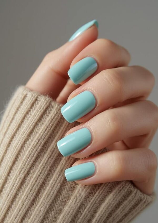 A hand with neatly manicured nails painted light blue, wearing a beige ribbed sweater, is shown against a neutral background.