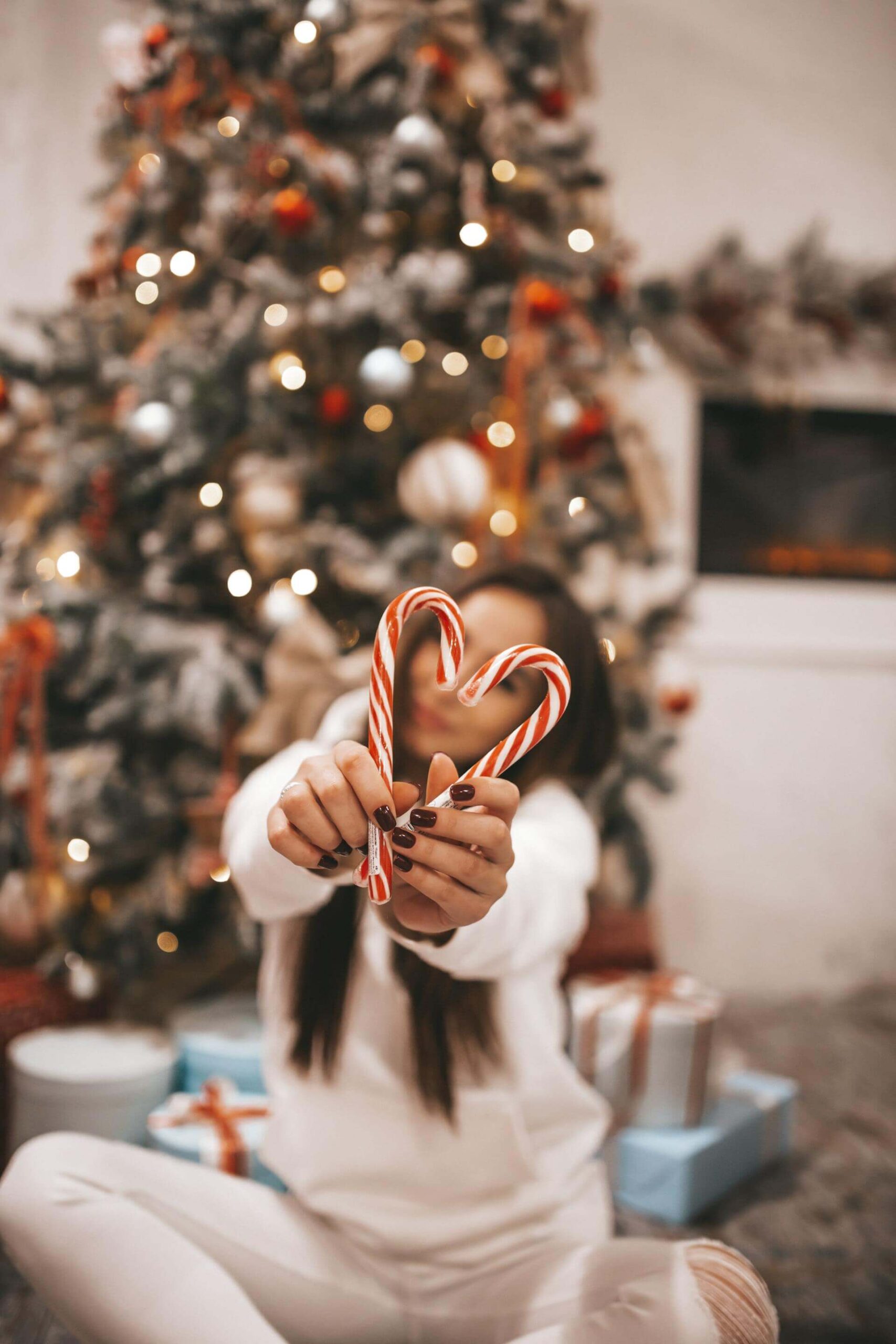 A person in white clothes holds two candy canes shaped like a heart in front of a decorated Christmas tree, with wrapped presents on the floor and warm lights in the background.