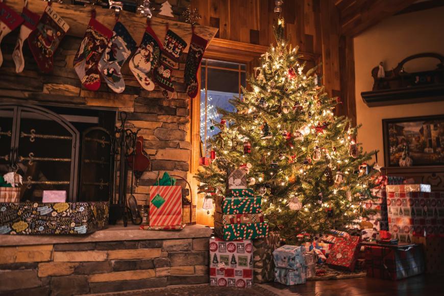 A decorated Christmas tree with glowing lights stands beside a stone fireplace hung with stockings. Wrapped gifts are piled under the tree and near the fireplace in a cozy, warmly lit living room.