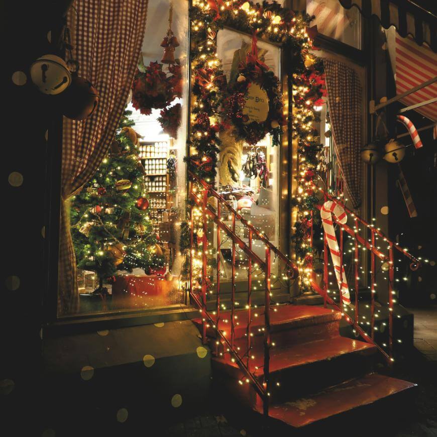 A warmly lit shop entrance decorated with Christmas lights, wreaths, and garlands. A red staircase leads up to the door. Inside, a Christmas tree and festive ornaments are visible through large windows.