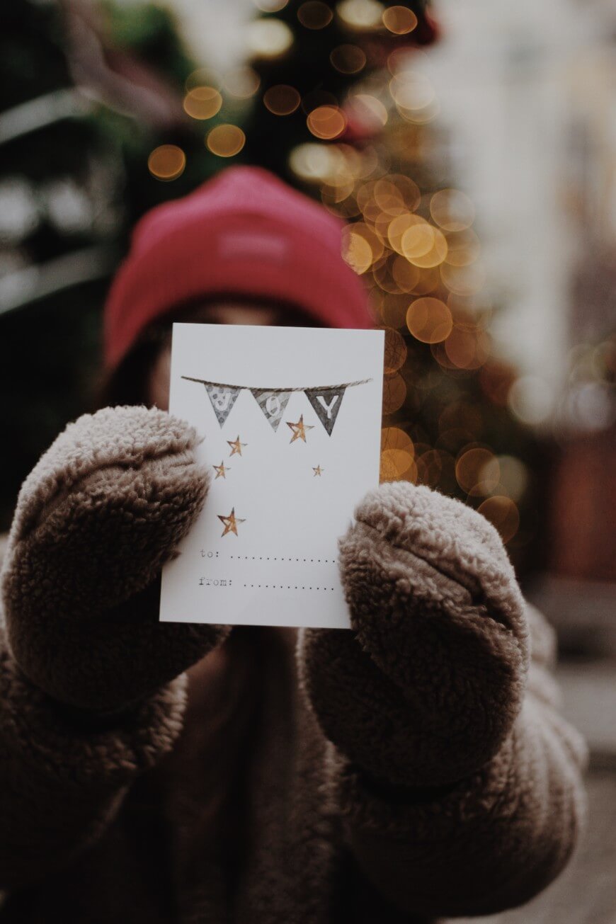 A person in a red beanie and brown mittens holds a festive card with stars and banners, with blurred holiday lights and a decorated tree in the background.