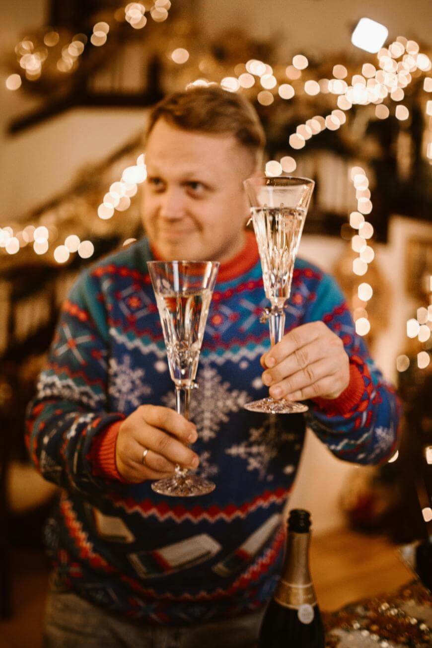 A person in a festive sweater smiles while holding up two crystal champagne glasses. Warm string lights and blurred holiday decorations create a cozy, celebratory atmosphere in the background.