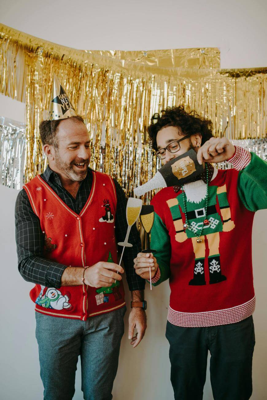 Two men wearing festive sweaters and party hats smile and hold fake champagne glasses and props, celebrating in front of a gold tinsel backdrop.
