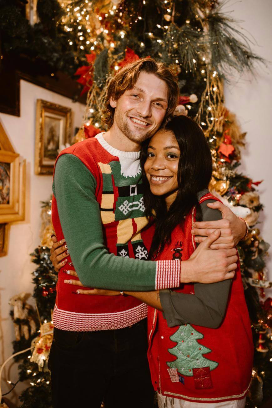 A smiling couple in festive sweaters hugs in front of a decorated Christmas tree with twinkling lights and ornaments. Both appear happy and cozy, surrounded by holiday decor.