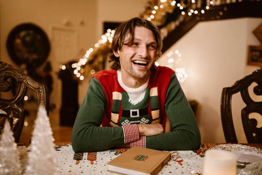 A man in a festive sweater smiles and laughs while sitting at a decorated table with holiday lights and ornaments in the background.
