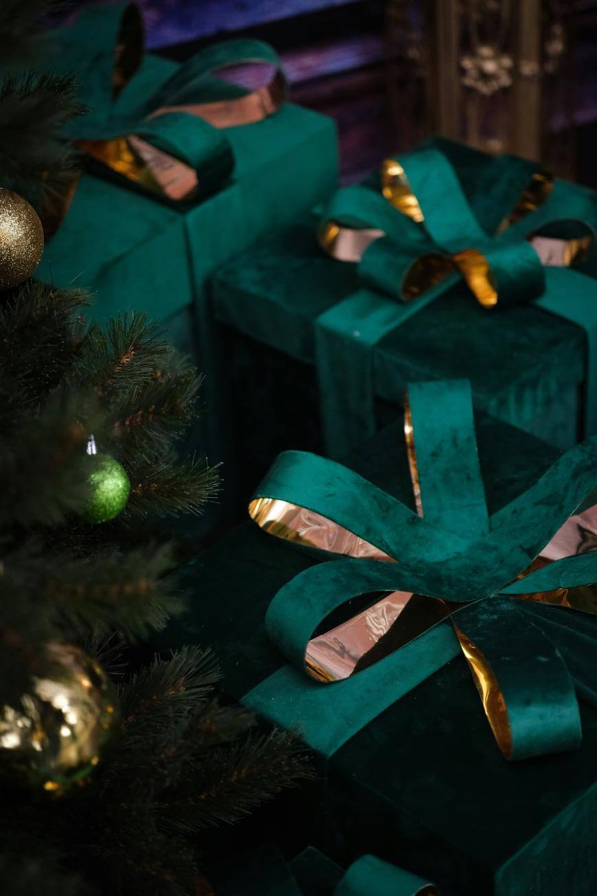 Large green gift boxes with shiny gold-accented ribbons are placed next to a decorated Christmas tree adorned with green and gold ornaments.