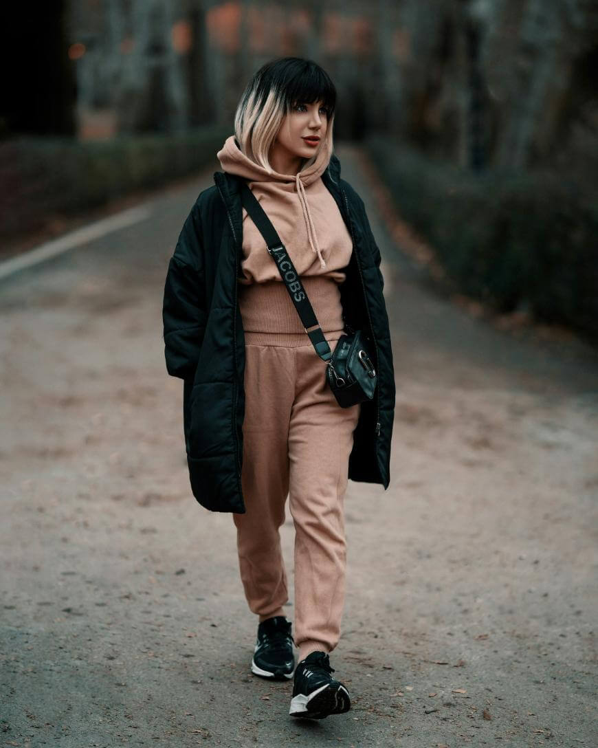 A person with shoulder-length hair and bangs walks on a path in a park, wearing a beige hoodie and sweatpants, a long black coat, black sneakers, and carrying a small black crossbody bag.