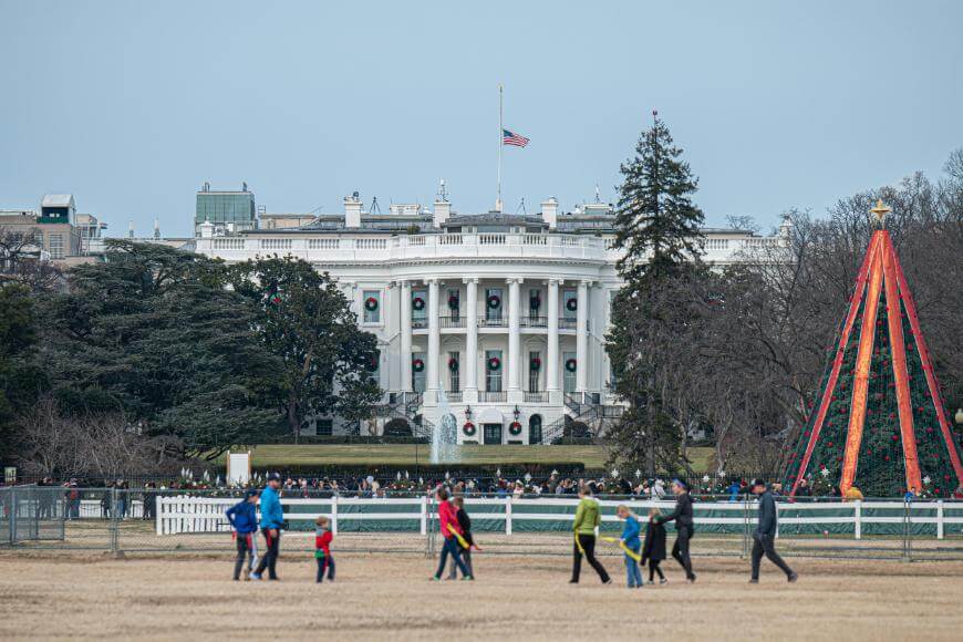 People walk on a grassy area in front of the White House, which is decorated with holiday wreaths. An American flag flies at half-mast, and a large Christmas tree is visible to the right.