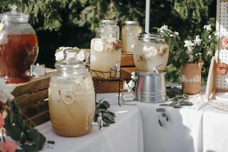 A table is set outdoors with large glass beverage dispensers filled with lemonade and iced tea, garnished with lemon slices. The table is decorated with flowers and greenery, creating a festive, rustic atmosphere.