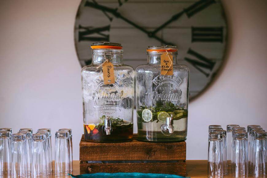 Two large glass drink dispensers with spigots sit on a wooden stand, each filled with different drinks and garnishes. Several empty glasses are arranged nearby, with a large clock in the background.