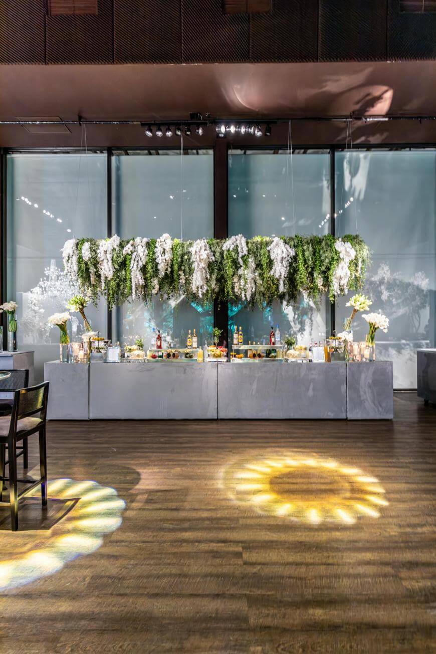 A modern bar setup features a large, suspended arrangement of cascading white flowers and greenery above a sleek counter with bottles and glassware. Soft lighting creates floral patterns on the wooden floor.