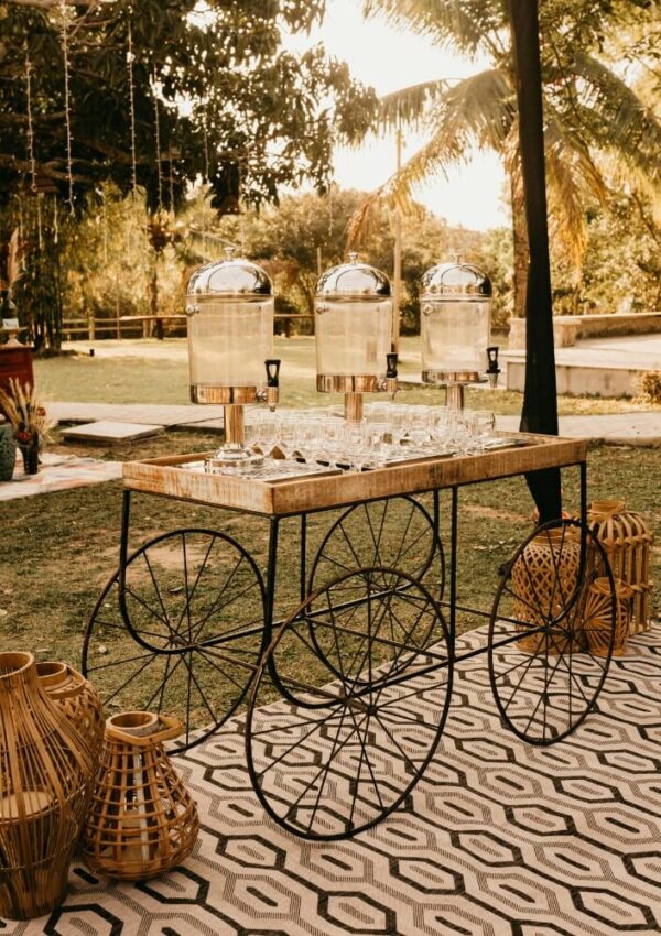 A rustic drink cart with glass dispensers and rows of glasses stands outdoors on a rug, surrounded by wicker lanterns and greenery, in warm, golden sunlight.