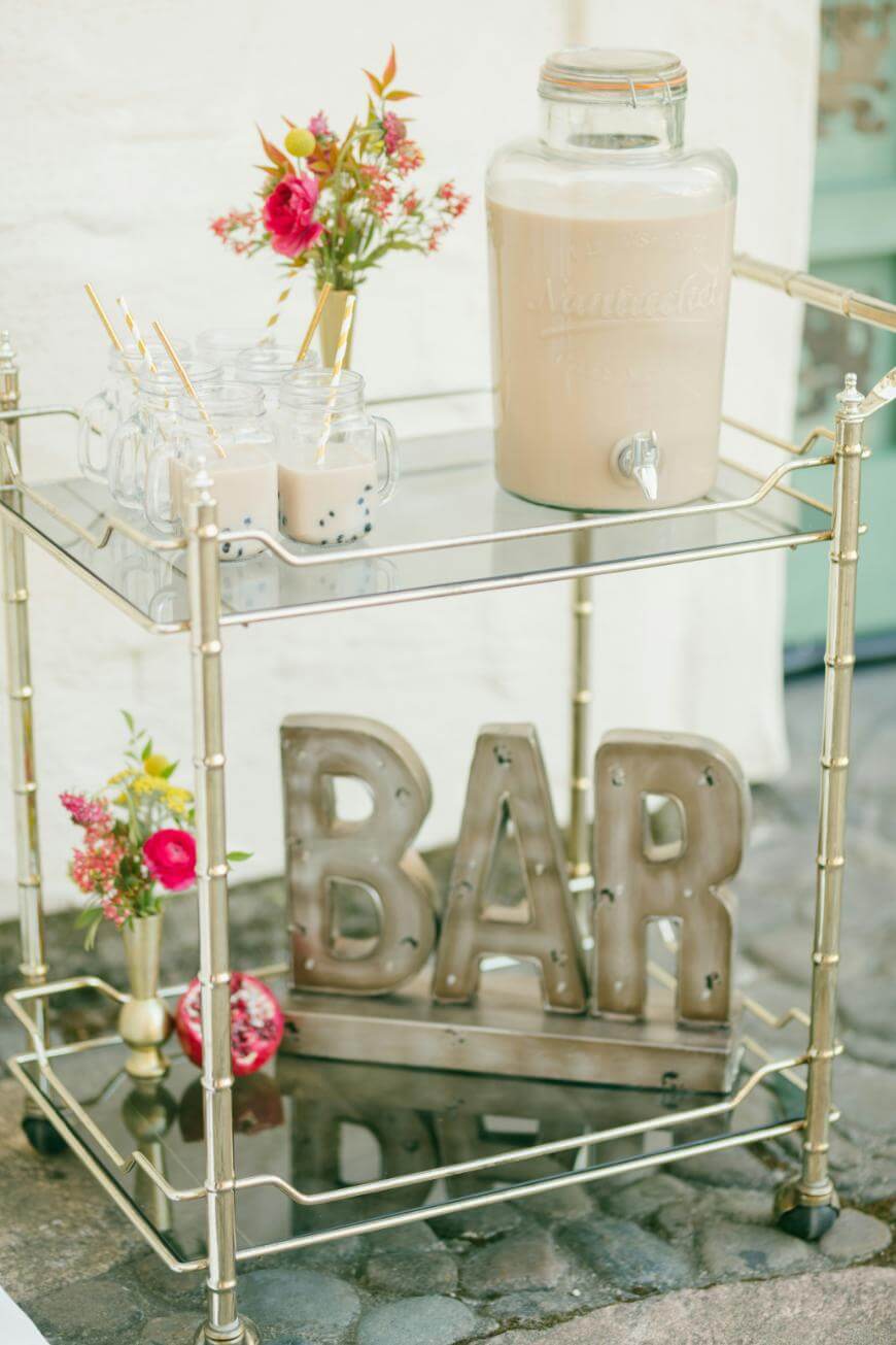 A gold bar cart with a glass jug of milk tea, four mason jar glasses with bubble tea and straws, flowers in small vases, and large metal letters spelling BAR on the lower shelf.