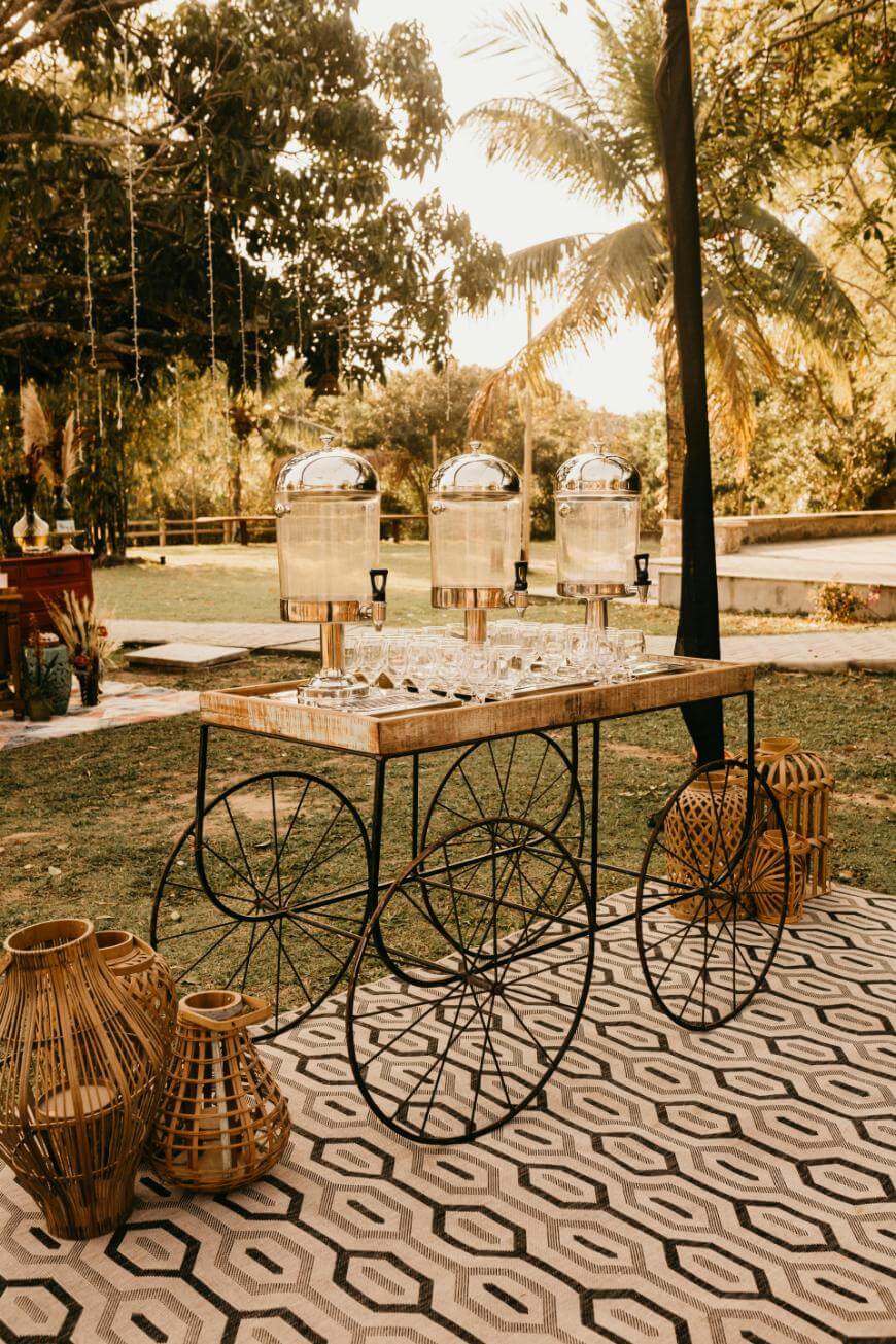 A decorative drink station with three glass dispensers and rows of glasses on a cart with large wheels, set outdoors on a geometric rug, surrounded by wicker lanterns and lush greenery.