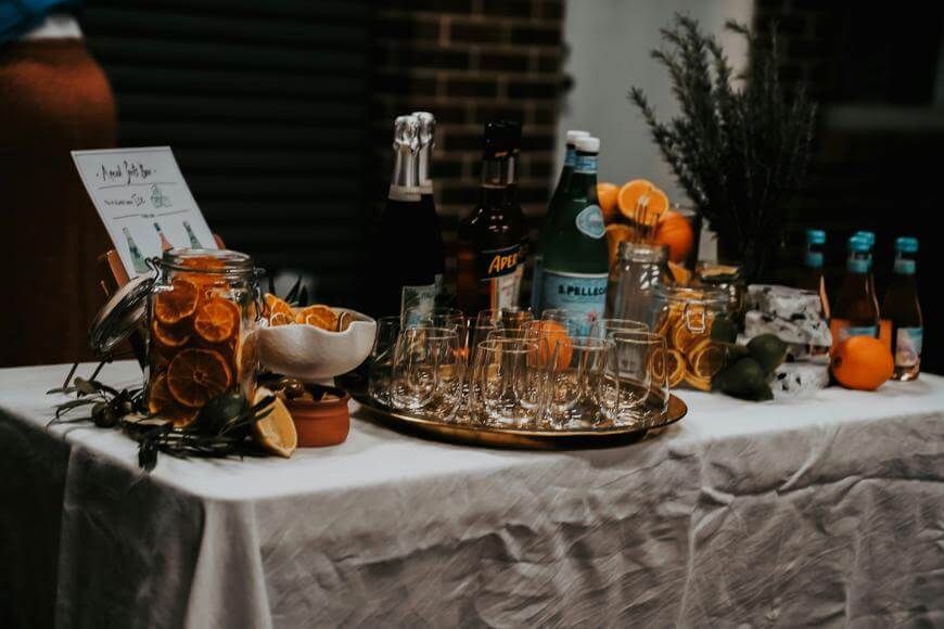 A table with a white tablecloth holds glass bottles, sliced citrus fruits, herbs, several empty glasses on a tray, and jars filled with dried orange slices, set up for making cocktails or drinks.