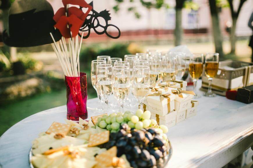 A table set outdoors with glasses of champagne, wrapped gift boxes, a plate of grapes and cheese, and a red vase holding fun photo props shaped like glasses, hats, and lips.