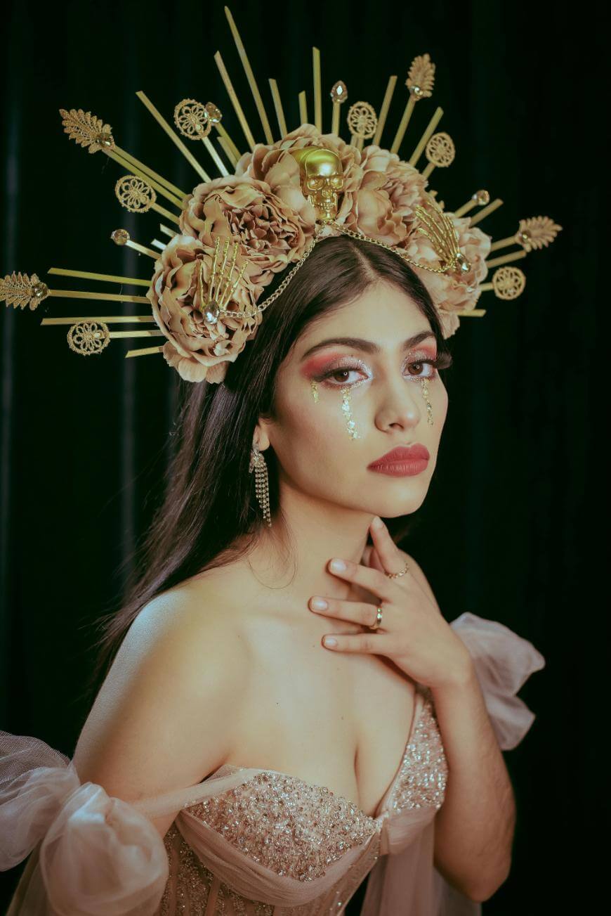 A woman in a jeweled, strapless dress poses against a dark background. She wears a gold floral and sunburst crown, with glitter tears on her cheeks and her hand resting on her collarbone.
