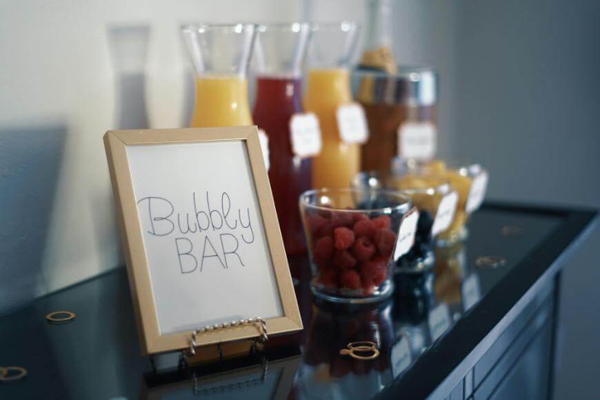 A Bubbly Bar sign sits on a counter next to glass pitchers filled with juice, a cocktail shaker, and bowls of fresh fruit including raspberries and blueberries, creating a stylish drink station.