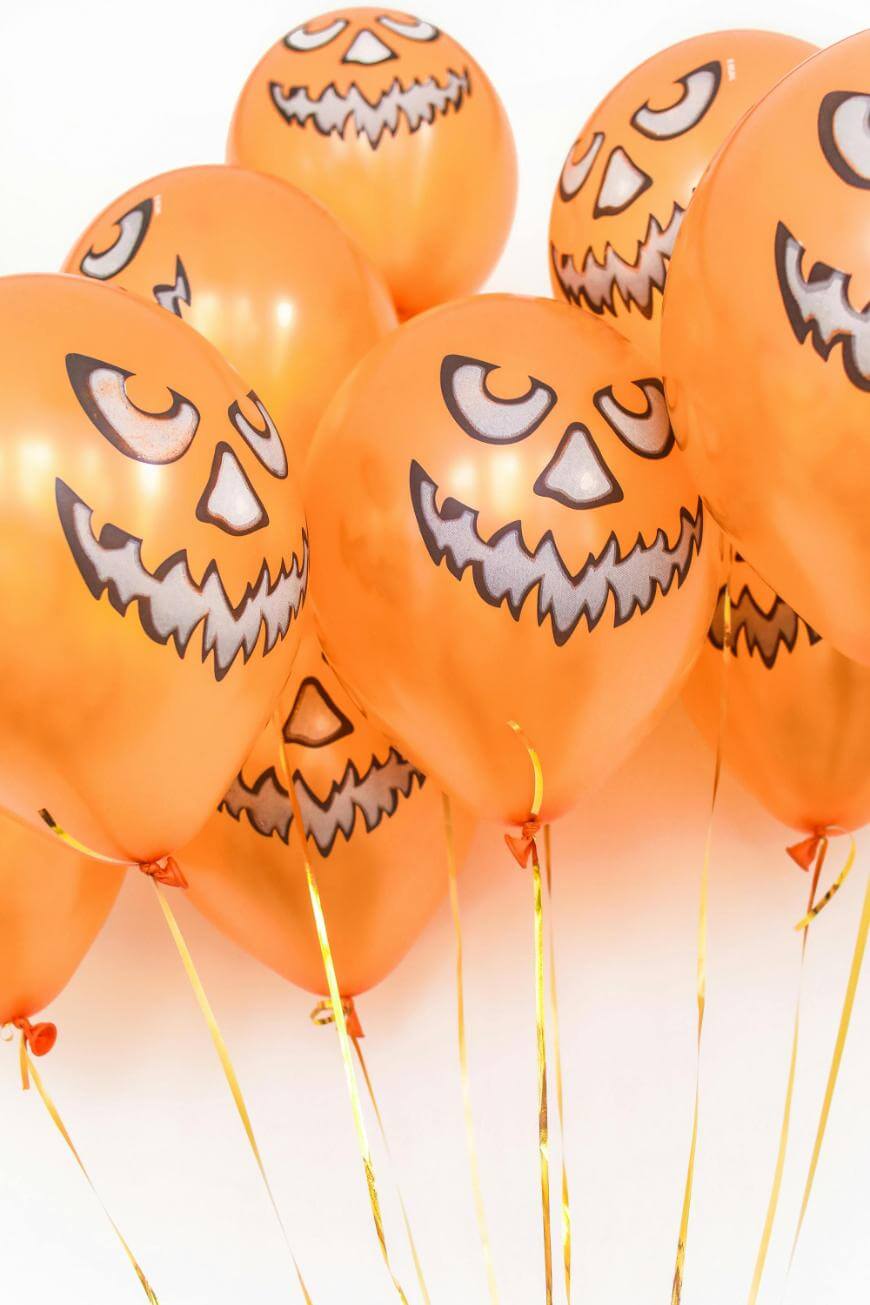 A group of orange balloons with black jack-o-lantern faces, arranged closely together against a white background, each balloon tied with a thin gold ribbon.
