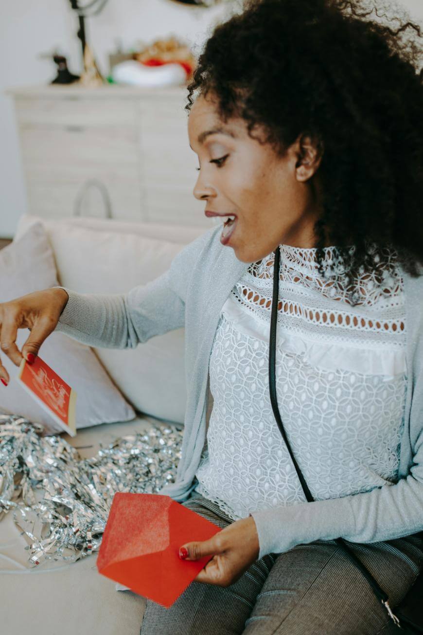 A woman with curly hair sits on a couch, smiling as she opens red envelopes. Silver tinsel is on her lap, and festive decorations can be seen in the background.