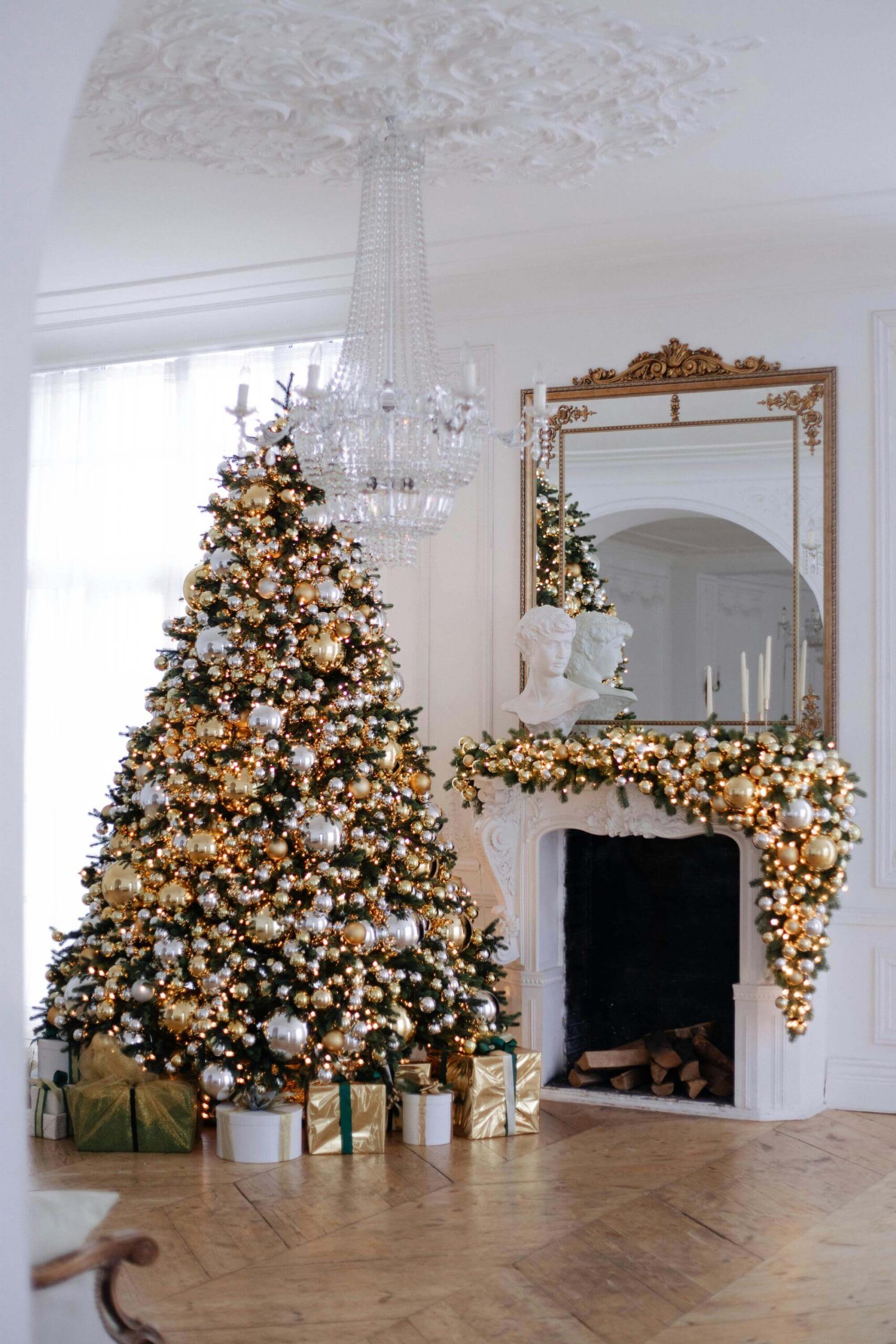 Elegant living room decorated for Christmas with a large, gold and white adorned Christmas tree, wrapped presents underneath, and a fireplace topped with garland, candles, and a large mirror above.