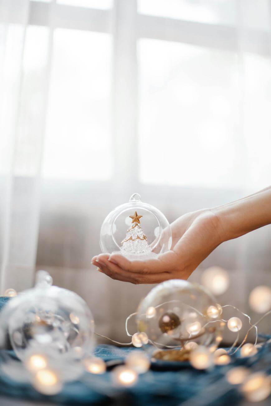 A hand holds a clear Christmas ornament containing a small white tree with a gold star. Two similar ornaments and string lights are on a blue surface. A bright window is in the background.