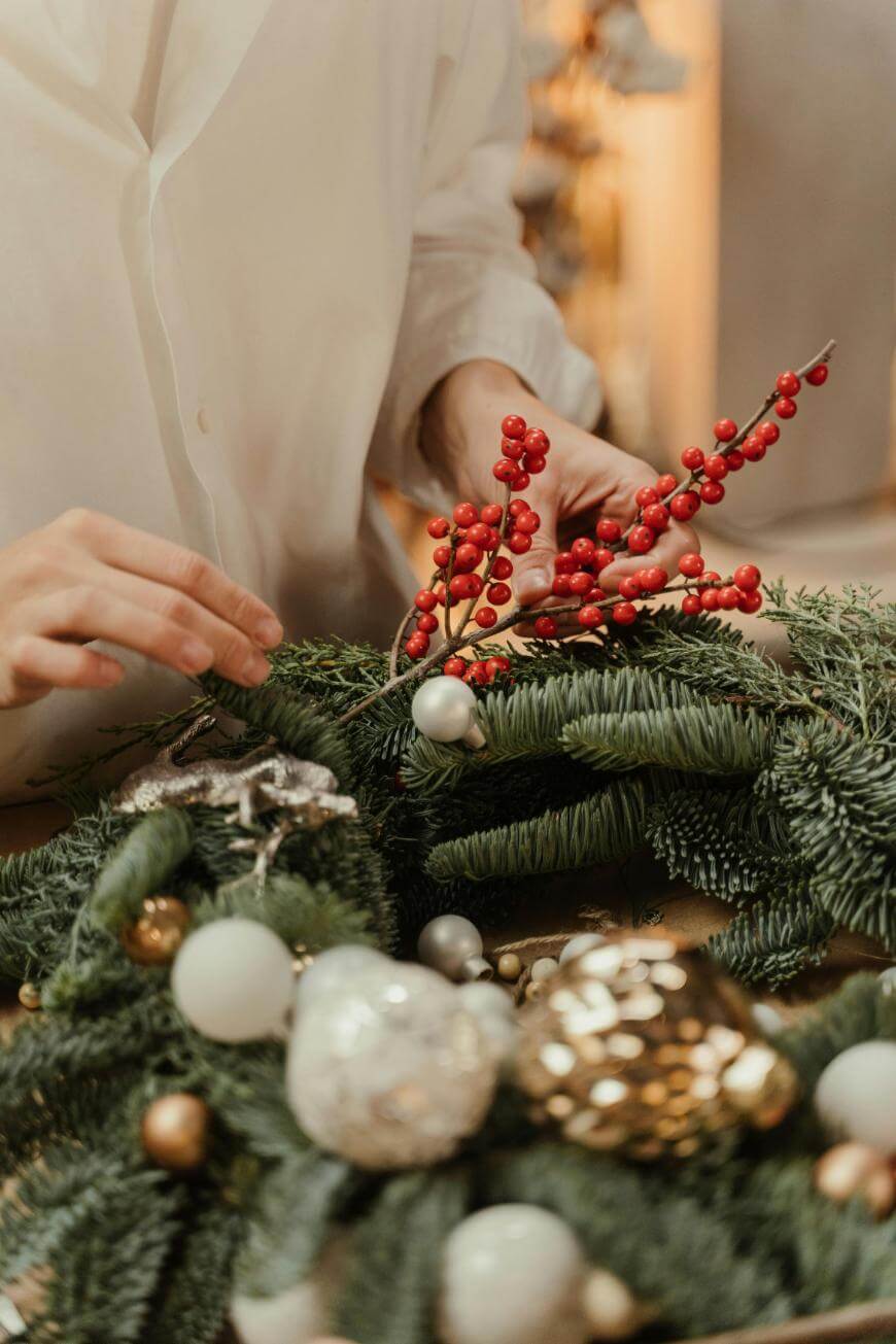 A person in a white shirt arranges red berries and evergreen branches on a holiday wreath decorated with ornaments, pine cones, and gold and white baubles.