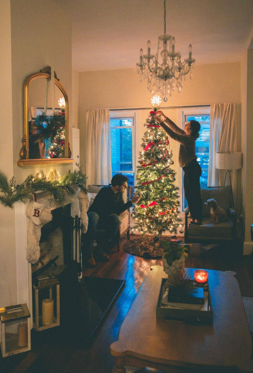 A cozy living room decorated for Christmas. One person places a star on top of a lit Christmas tree while another sits nearby watching. A dog stands by the tree, and stockings hang by the fireplace. Candles and lights add warmth.