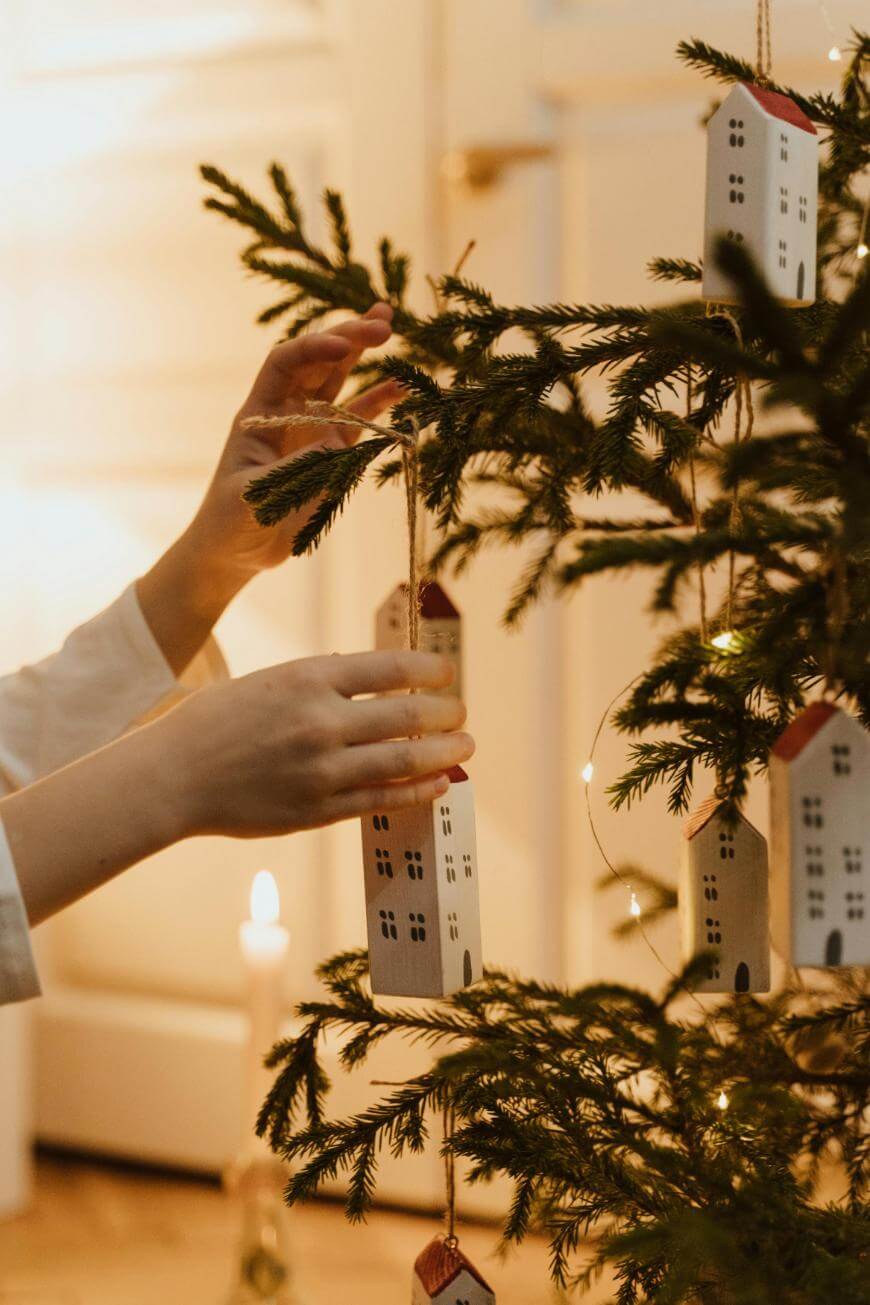 Hands hang small house-shaped ornaments on a decorated Christmas tree with warm lights, with a lit candle glowing softly in the background.