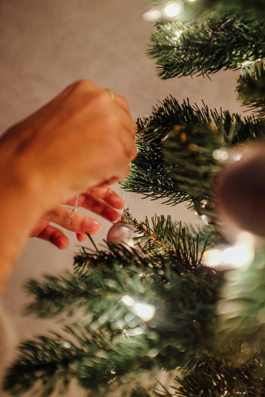 A hand hangs a silver ornament on a Christmas tree decorated with warm white lights, creating a festive and cozy atmosphere.