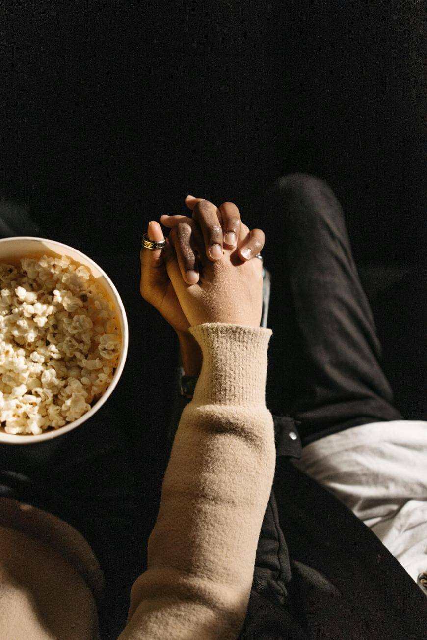 Two people sit close together, holding hands, with a large bowl of popcorn in one person’s lap. Both are wearing cozy, light-colored sweaters. The scene suggests intimacy and relaxation, possibly watching a movie.