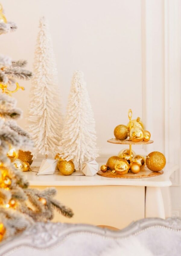 A festive scene with white faux Christmas trees, gold ornaments on a tiered tray, and a decorated white Christmas tree with warm lights, set against a light-colored wall.