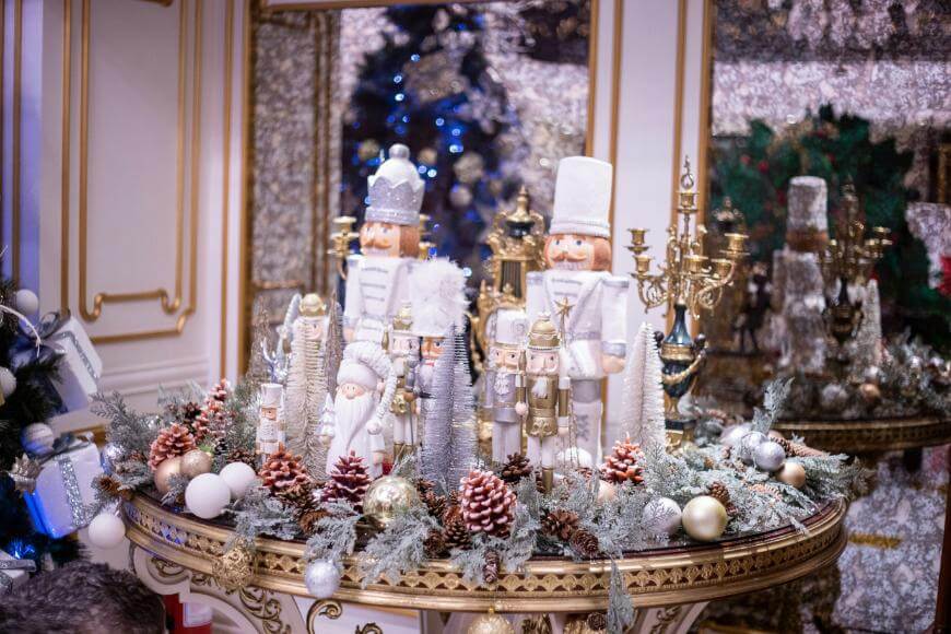 A festive table centerpiece features nutcracker figurines, miniature Christmas trees, pinecones, ornaments, and greenery, all arranged on a decorative round table with a gold-trimmed mirror and holiday decor in the background.