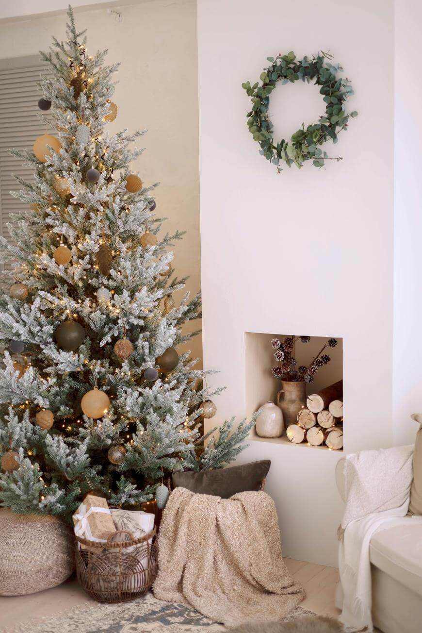 A decorated Christmas tree with gold ornaments and lights stands beside a white fireplace filled with logs. A green wreath hangs above the fireplace, and a cozy chair with a blanket and pillow is nearby.