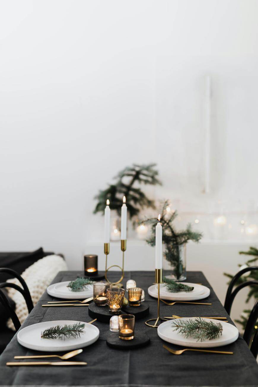 A modern dining table set for four, decorated with white plates, gold cutlery, candles, and pine sprigs on each plate. The table has a black tablecloth and festive greenery as centerpiece accents.