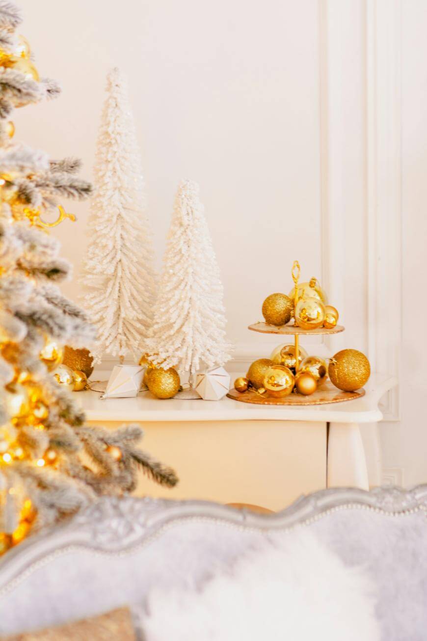 A festive scene with white flocked Christmas trees, gold ornaments, and a tiered tray of gold decorations on a white table. A frosted Christmas tree with warm lights is partially visible in the foreground.