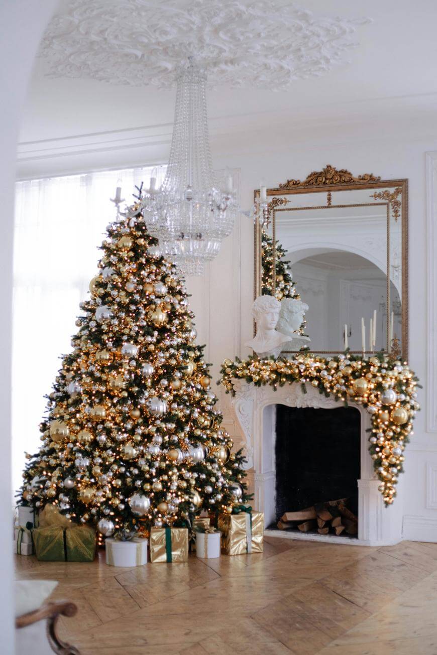 A lavishly decorated Christmas tree with gold and white ornaments stands beside a white fireplace adorned with gold garland, candles, and a bust, with wrapped gifts below, in an elegant, light-filled room.