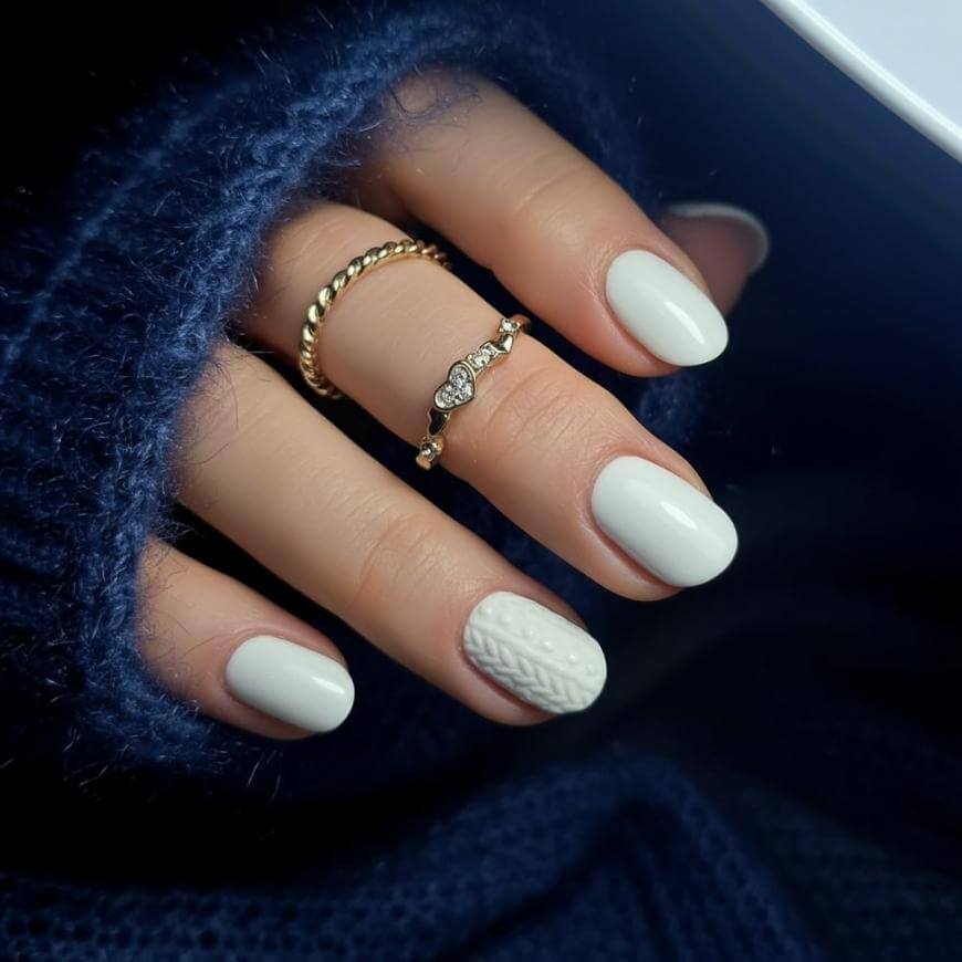 A hand with short, oval-shaped nails painted white, featuring a knitted 3D pattern on the ring fingernail. The hand wears two gold rings and is partially wrapped in a dark blue, fuzzy sweater.