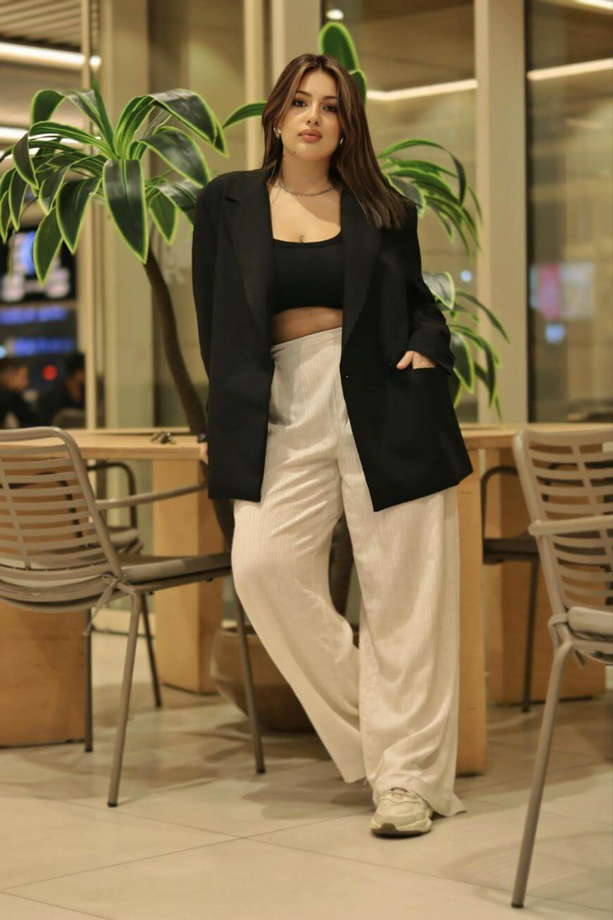 A woman stands indoors by a table and chairs, wearing a black blazer over a black crop top, white wide-legged pants, and white sneakers, with a green plant in the background.