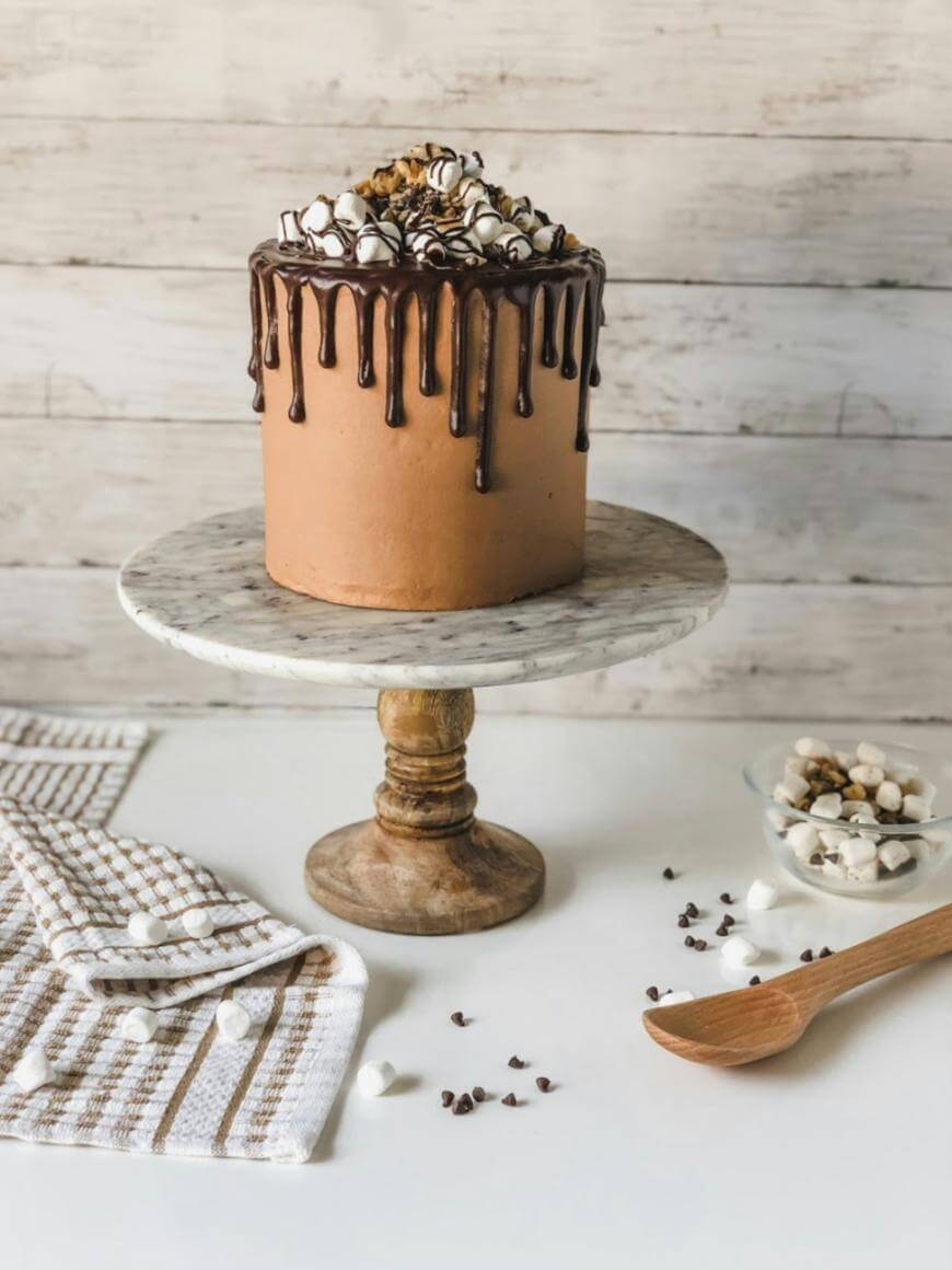 A chocolate drip cake topped with marshmallows, chocolate chips, and nuts sits on a marble and wood cake stand. Nearby are a wooden spoon, a bowl of toppings, and a checkered kitchen towel.