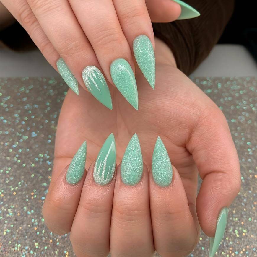 Hands with long, pointed acrylic nails painted in mint green. Some nails have a glittery finish, while others are glossy with a subtle white accent design. The background is sparkly and complements the nail color.