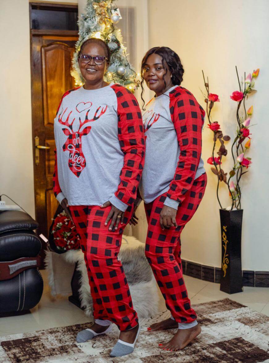 Two women stand indoors in matching festive pajamas with red and black checkered pants and gray tops featuring reindeer designs. A decorated Christmas tree and flower arrangements are visible in the background.