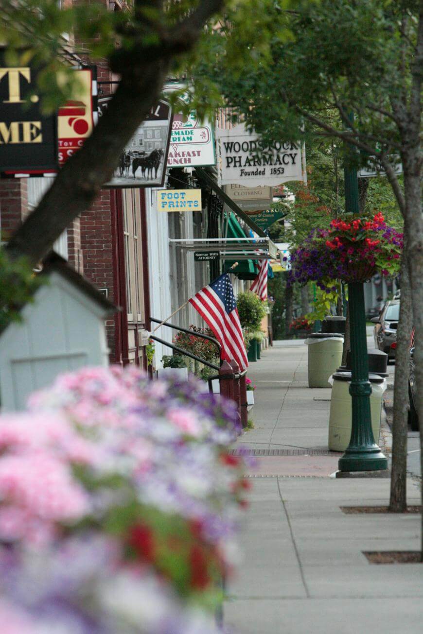 A small-town sidewalk lined with storefronts, American flags, hanging flower baskets, and blooming flowers in the foreground; signs for a pharmacy and breakfast place are visible.